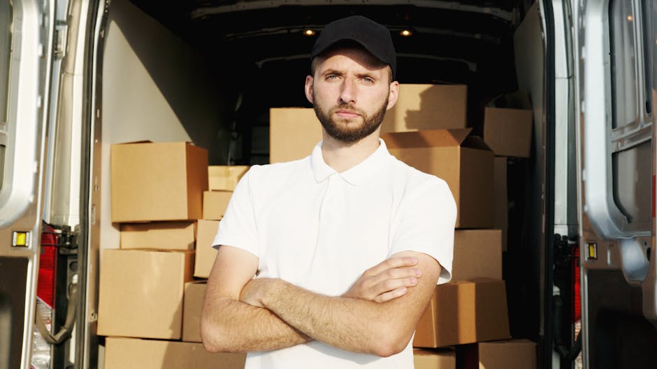 A young man with a beard, wearing a black cap and a white polo shirt, stands inside the back of a large moving van filled with stacked cardboard boxes and packing materials. The boxes are of various sizes and appear to be securely packed, some wrapped in plastic or placed on top of foam padding. The van's interior is well-lit, highlighting the orderly arrangement of the boxes, and the man is standing with his arms crossed, looking directly at the camera with a neutral expression. Outside the open rear doors, part of the pavement and the exterior of the van are visible, indicating an outdoor home relocation scene. This image accurately depicts the process of packing and loading items for a house move, aligning with services offered by Man and Van Finsbury Park for furniture transport and moving logistics during house removals.