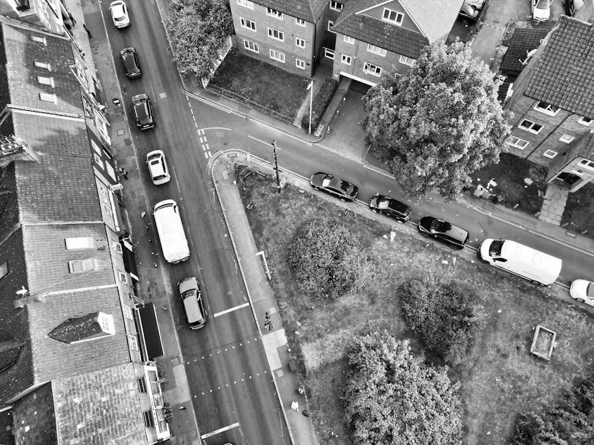A street scene in Finsbury Park during the daytime, featuring a church with a tall clock tower in the background surrounded by green leafy trees. Parked cars line both sides of the street, with some vehicles waiting at a traffic light. In the foreground, a man from Man and Van Finsbury Park, a professional removals company, is engaged in a home relocation task, carrying a cardboard box wrapped in plastic, with other packed boxes and moving blankets nearby. The area appears calm with clear skies overhead, and the scene captures the process of furniture transport and packing and moving activities typical of house removals in the Finsbury Park area.