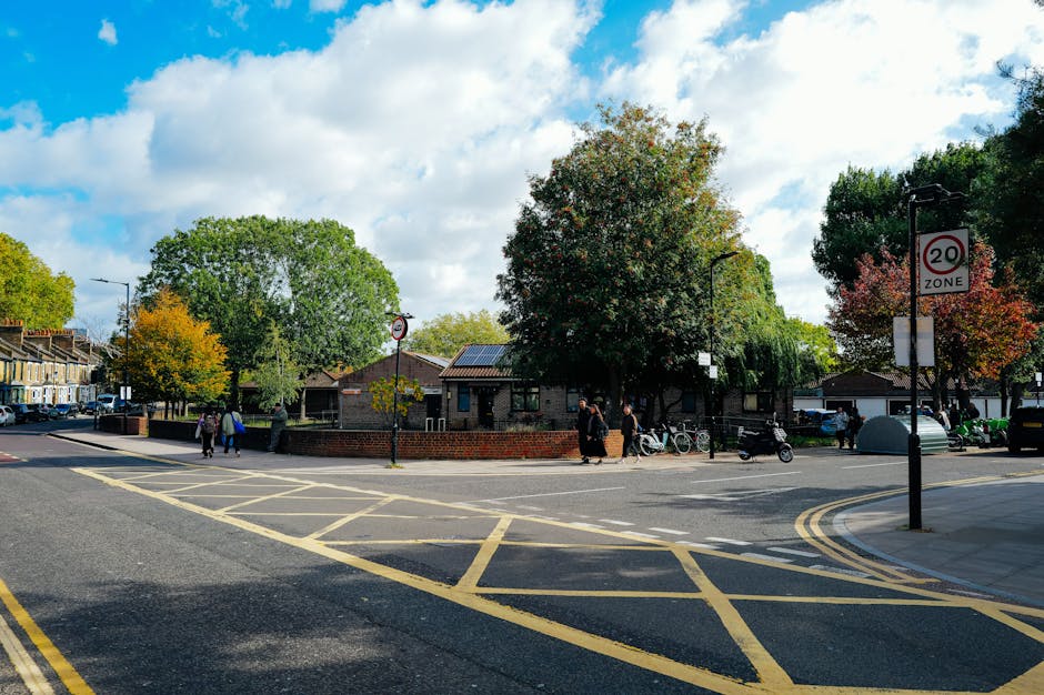 A street scene on Stroud Green Road in Finsbury Park, showing a small public green space surrounded by a low brick wall, with several trees displaying autumn foliage. In the foreground, there are yellow box junction markings and a double yellow line along the edge of the road. Pedestrians are walking along the pavement, some crossing the street near a pedestrian crossing sign, while others stand beside parked bicycles and motorbikes. To the right, a black lamppost displays a 20 mph speed limit zone sign. Behind the green space, residential buildings with sloped roofs and solar panels are visible under a partly cloudy sky. The scene is well-lit with natural daylight, highlighting the foliage and urban environment, reflecting a typical residential neighbourhood where house removals or furniture transport could take place, often involving loading and unloading nearby.