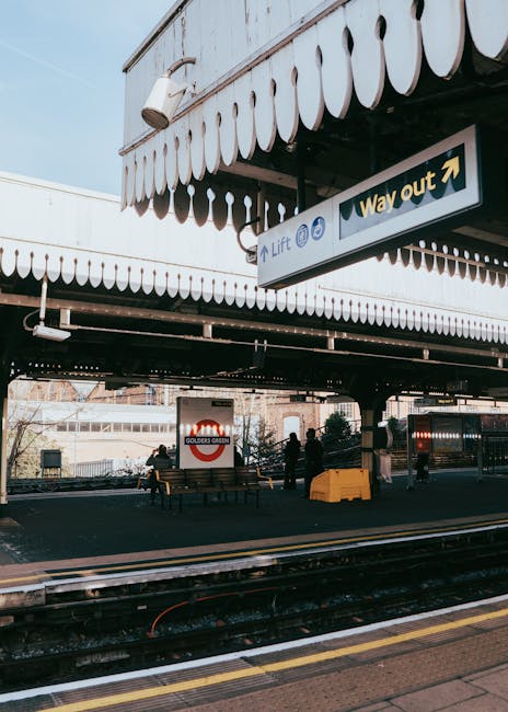 The image shows a train station platform beneath an overhanging building structure with a decorative scalloped edge. There is a sign indicating the way out and directions to lifts, hanging beneath the eaves of the building. Several parked benches are visible on the platform, and a few people are standing nearby, possibly waiting for a train or during a house move. In the background, railway tracks are visible with some equipment on the platform, which is surfaced with paving stones. The scene suggests a busy transit environment relevant to relocation and moving logistics, with a focus on transportation facilities. Man and Van Finsbury Park's removals service could utilize such images to illustrate efficient travel and transfer processes related to home relocations, with evidence of a well-organized transit hub suited for furniture transport and packing and moving tasks.