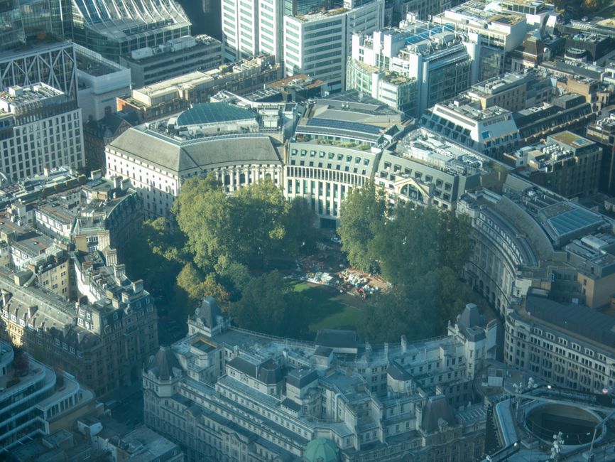 An aerial view of a cityscape showing a large circular park surrounded by historic and modern buildings, with tall office towers and residential structures densely packed around the park. The park features green lawns and several mature trees, with some construction or moving materials and vehicles visible within. The image captures the urban environment during daylight with clear lighting and shadows cast by the buildings, illustrating a typical scene of city infrastructure supporting home relocations and moving logistics within the area, as seen in the context of Finsbury Park Station N4 and services offered by Man and Van Finsbury Park.