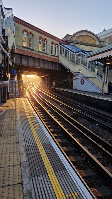 The image shows a train station platform beneath an overhanging building structure with a decorative scalloped edge. There is a sign indicating the way out and directions to lifts, hanging beneath the eaves of the building. Several parked benches are visible on the platform, and a few people are standing nearby, possibly waiting for a train or during a house move. In the background, railway tracks are visible with some equipment on the platform, which is surfaced with paving stones. The scene suggests a busy transit environment relevant to relocation and moving logistics, with a focus on transportation facilities. Man and Van Finsbury Park's removals service could utilize such images to illustrate efficient travel and transfer processes related to home relocations, with evidence of a well-organized transit hub suited for furniture transport and packing and moving tasks.